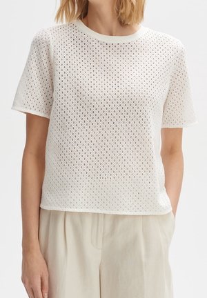 Frau trägt ein weißes, kurzärmeliges Oberteil aus Häkelstrick mit rundem Ausschnitt, kombiniert mit beigen hoch taillierten Hosen, die Hände in den Taschen.