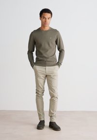 Olive zelena srajca z dolgimi rokavi, bež slim-fit hlače in temno rjavi gležnjarji, stoji na svetlo bež tleh ob svetli steni.