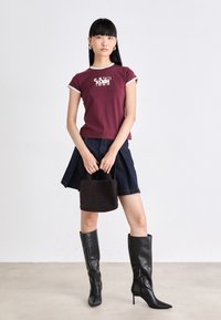 Maroon short-sleeve top with contrasting trim, denim skirt, black knee-high pointed boots, and a small dark textured handbag.