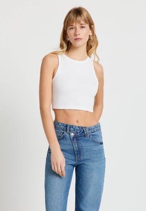 Giovane donna con capelli chiari che indossa un top bianco senza maniche e jeans blu a vita alta, su sfondo bianco uniforme.