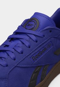 Μπλε αθλητικό παπούτσι Reebok με υφασμένες κορδόνια, μαύρες λωρίδες λογότυπου, όνομα της μάρκας και καφέ σόλα ορατή.
