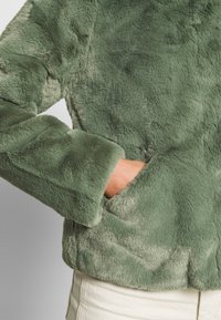 Personne portant une veste en fausse fourrure verte douce, main glissée dans la poche latérale, associée à un pantalon clair.