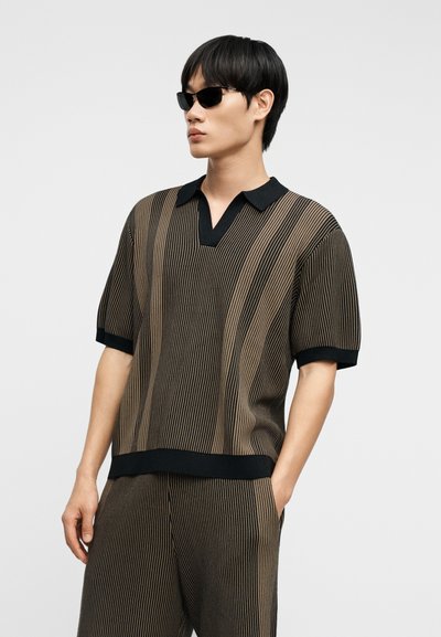 Jeune homme portant des lunettes de soleil noires et un polo à manches courtes rayé verticalement marron et noir avec un pantalon assorti, debout avec une main dans la poche.