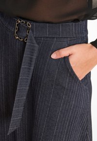 Hand in der Tasche von dunkelblauen Nadelstreifen-Hosen mit Fischgrätmuster, ausgestattet mit einem passenden Stoffgürtel und einer dekorativen quadratischen Schnalle.