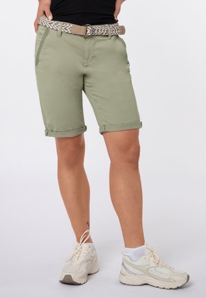 Person trägt hellgrüne hochgekrempelte Shorts mit einem gewebten Gürtel, weiße Sneakers und ein schwarzes Top, steht mit den Händen in der Hüfte.