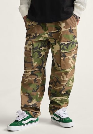 Pantalones cargo de camuflaje en tonos de verde y marrón, con múltiples bolsillos, un ajuste relajado y fabricados con tejido duradero. Combinados con zapatillas verdes.