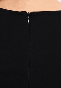 Haut en tissu noir côtelé avec une fermeture éclair discrète au dos. L'encolure est large et la coupe est ajustée.