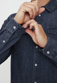 Camisa de mezclilla en azul oscuro, con cuello, dos bolsillos frontales y puños abotonados. La tela tiene una ligera textura, con botones de plástico.