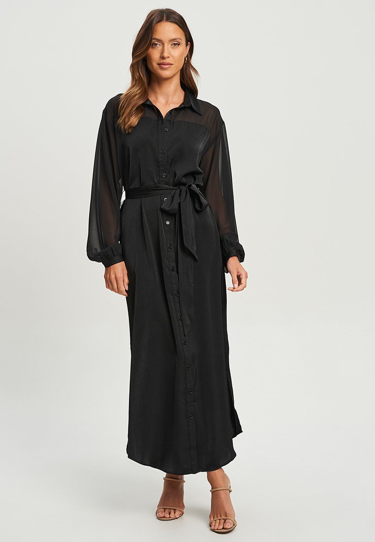 Robe noire à manches longues avec un devant à boutons, ceinture nouée à la taille et une silhouette fluide. Manches transparentes et texture de tissu lisse.