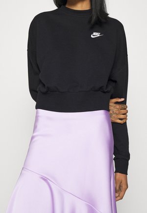 Personne portant un sweat-shirt Nike noir et une jupe en satin violet clair, une main aux doigts tatoués reposant sur le bras opposé.