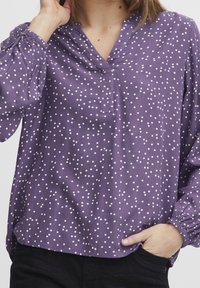 Lila blus med vita prickar, V-ringning, långa ärmar med elastiska ärmslut och en lös passform. Mjuk tygtextur och avslappnad design.