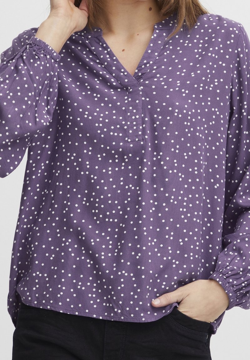Lila blus med vita prickar, V-ringning, långa ärmar med elastiska ärmslut och en lös passform. Mjuk tygtextur och avslappnad design.