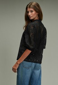 Schwarzes Spitzenoberteil mit Puffärmel und hohem Hals, mit floralen Mustern und transparenter Textur, kombiniert mit hellblauen, weiten Jeans.