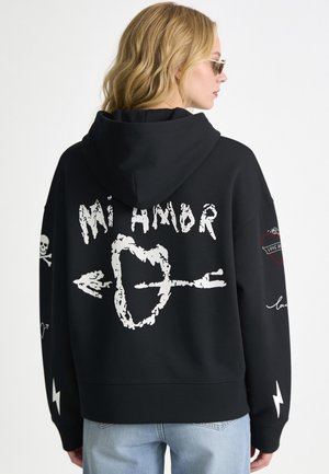 Donna bionda che indossa una felpa nera con cappuccio con cuore bianco trafitto da una freccia e testo "Mi Amor" sulla schiena, oltre a disegni di teschi e fulmini sulle maniche.