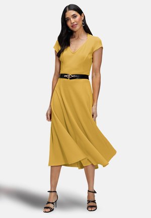 Yellow, short-sleeve dress with a V-neck and flowing skirt, cinched at the waist by a black belt with a gold accent. Worn with black strappy sandals.