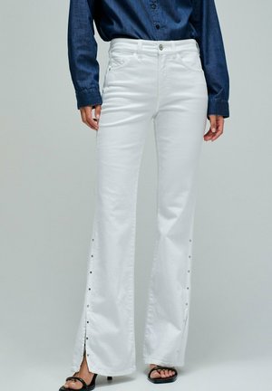 Mujer con pantalones blancos de pierna ancha y cintura alta con cierres laterales, combinados con una camisa de manga larga azul oscuro y sandalias negras de tacón.