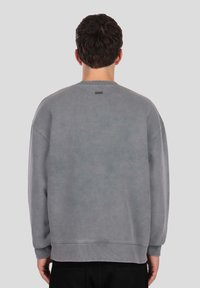 Sivá mikina s voľným strihom, okrúhlym výstrihom, rebrovanými manžetami a malou značkovou nášivkou na hornej časti chrbta. Mäkká textúra.