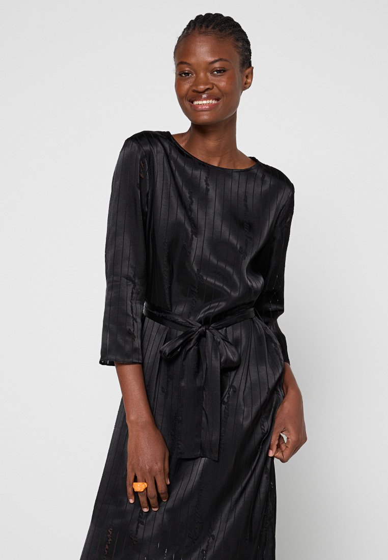 Černé hedvábné šaty s volnějším střihem, třičtvrtečním rukávem, strukturovanými vertikálními pruhy a uvázaným pasem. Obsahují oranžový výrazný prsten.