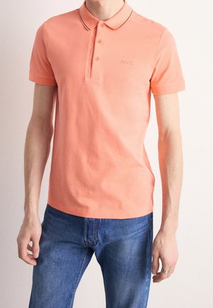 Korallfarbenes Poloshirt mit dreiknöpfiger Knopfleiste und kontrastierendem Kragenbesatz, kombiniert mit blauen Denim-Jeans. Verfügt über ein dezentes Logo auf der Brust.