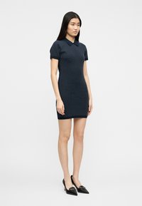 Γυναίκα που στέκεται με φόρεμα σε ναυτικό μπλε χρώμα, με κοντά μανίκια, μήκος μέχρι τα γόνατα και λευκές πουά, φορώντας μαύρες γόβες με μυτερή μύτη σε απλό φόντο.