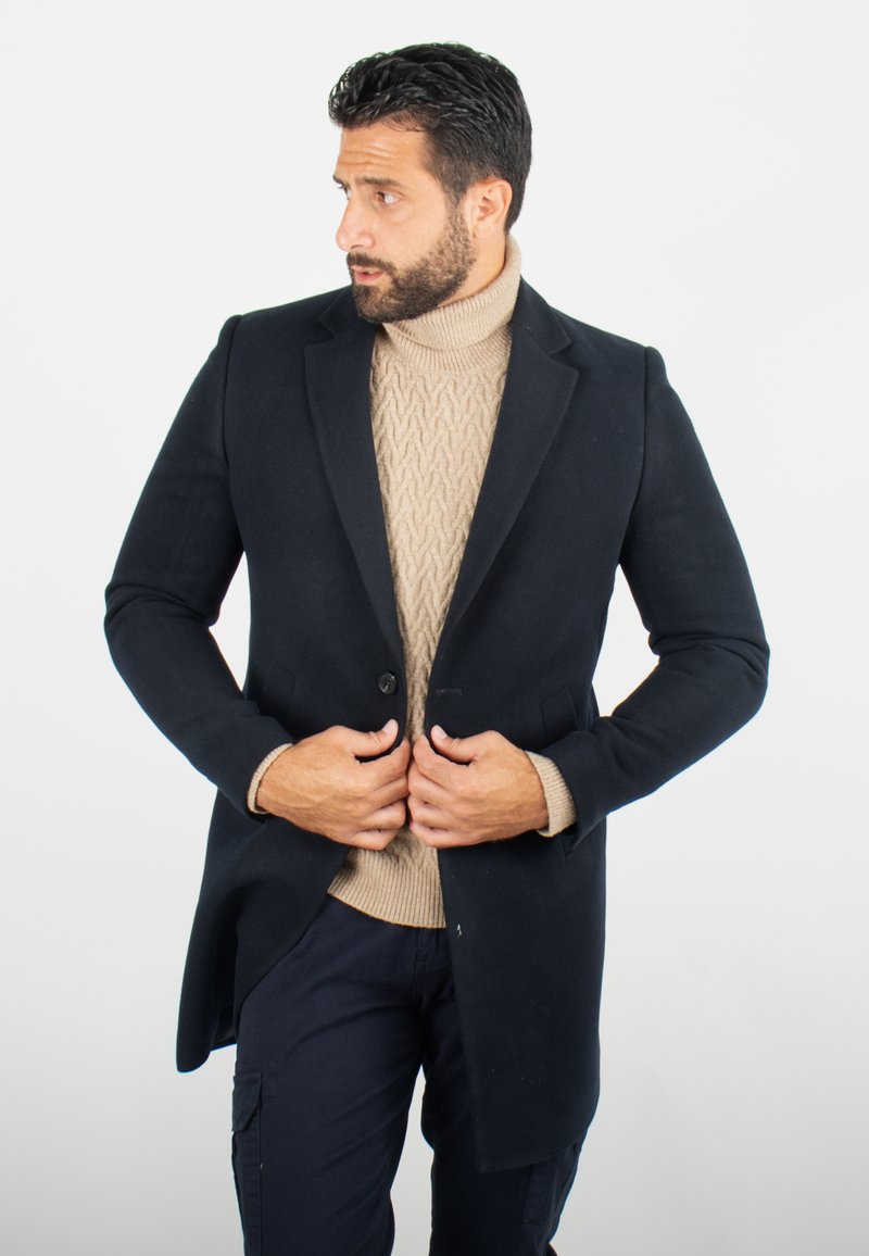 Manteau en mélange de laine navy avec une coupe ajustée, fermeture à un bouton, et deux poches avant, porté sur un pull en maille beige à col roulé.