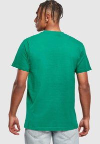 Grøn bomulds t-shirt med rund hals, korte ærmer og et enkelt design, vist fra ryggen med en ensfarvet tekstur. Denimjeans kan også ses.