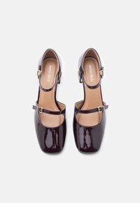 Fényes sötétlila Mary Jane sarkú cipők párja, szögletes orral, bokapántokkal és alacsony tömbsarkakkal, fehér háttér előtt, felülről nézve.