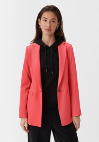 Jasnoróżowy żakiet z pojedynczym zapięciem na guzik, klapami i kieszenią na zamek, nałożony na czarny hoodie. Gładka tekstura materiału.