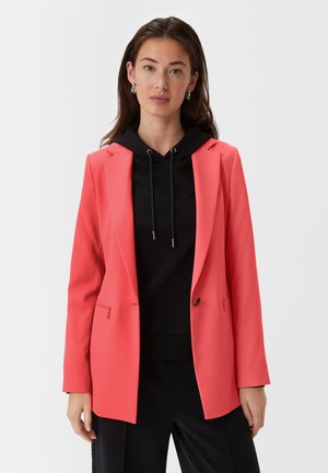 Jasnoróżowy żakiet z pojedynczym zapięciem na guzik, klapami i kieszenią na zamek, nałożony na czarny hoodie. Gładka tekstura materiału.