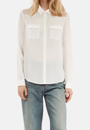 Biała koszula na guziki z koronkowymi panelami na ramionach i dwoma kieszeniami na piersiach, noszona z jasnoniebieskimi dżinsami.