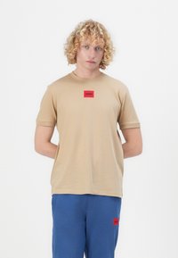 Beige bavlněné tričko s krátkým rukávem a kulatým výstřihem, na hrudi s červeným obdélníkovým nášivkou s nápisem "HUGO".