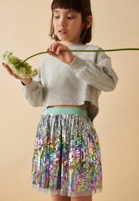 Felpa grigia corta, gonna plissettata con paillettes arcobaleno e vita azzurra. Bambino che tiene un fiore su uno sfondo beige.