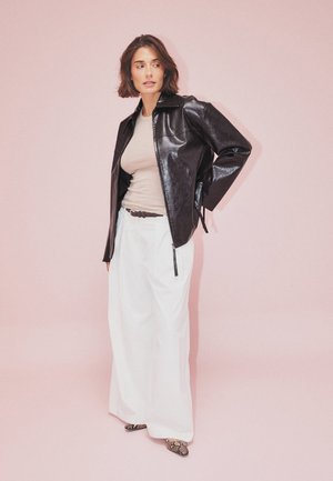 Veste en cuir noir avec fermeture éclair, haut beige, pantalon blanc ample et chaussures à motifs. Sur un fond rose pâle.