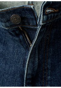 Gros plan d'un jean en denim bleu foncé avec une braguette partiellement ouverte, bouton métallique visible et coutures orange.