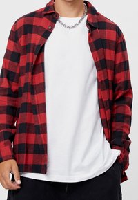 Rood-zwart geruit flanellen overhemd met een ontspannen pasvorm, knoopsluiting aan de voorkant en lange mouwen, gedragen over een witte T-shirt met ronde hals. Zilveren kettingaccent.