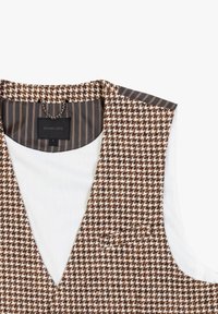 Gilet a motivo houndstooth marrone e bianco con un interno bianco e tessuto a strisce sul collo. Presenta una piccola tasca sul davanti.
