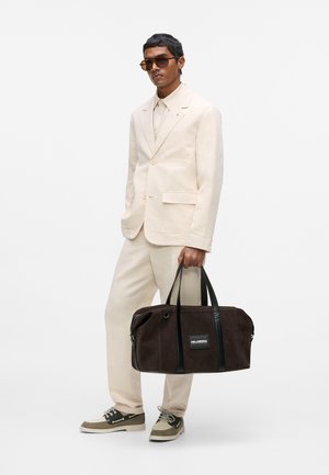 Uomo che indossa un completo color crema e occhiali da sole, tiene una grande borsa da viaggio marrone scuro con doppi manici su uno sfondo bianco uniforme.