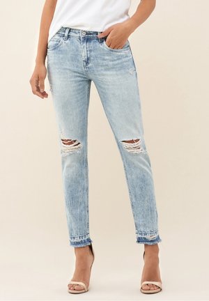 Vrouw draagt lichtblauwe gescheurde skinny jeans met rafelige zomen en beige open teen hakken, één hand in de zak, tegen een neutrale achtergrond.