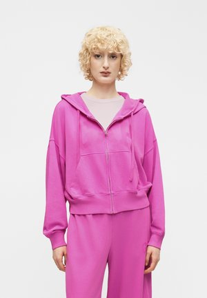 Persoon met kort, krullend blond haar draagt een felroze hoodie met ritssluiting en bijpassende wijd uitlopende broek tegen een effen witte achtergrond.