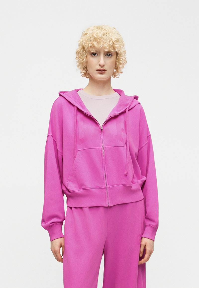 Personne aux cheveux blonds courts et bouclés portant un sweat à capuche zippé rose vif et un pantalon large assorti sur un fond blanc uni.