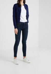 Marinblå stickad cardigan, vit skjorta med flerfärgade mönster, mörka slim fit jeans och vita sneakers. Slät textur, figursydd design.
