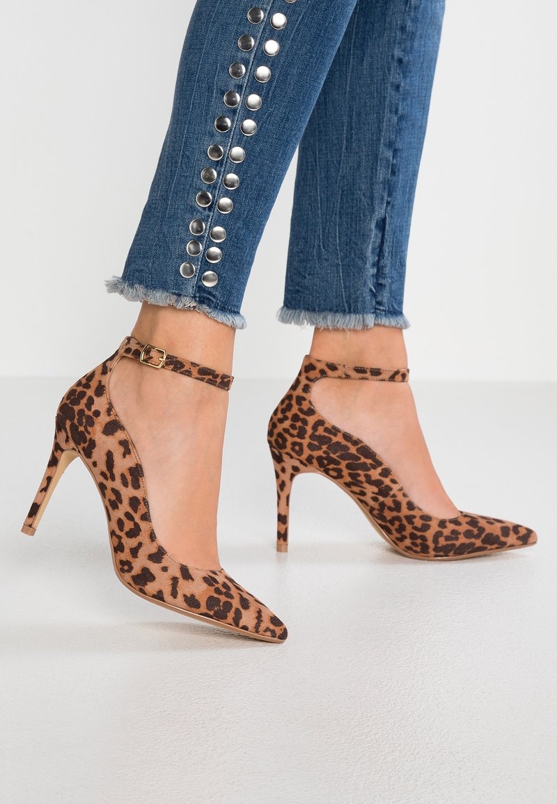 Personne portant un jean bleu avec des clous métalliques et des ourlets effilochés ainsi que des escarpins à bout pointu imprimé léopard avec brides à la cheville sur un sol blanc.