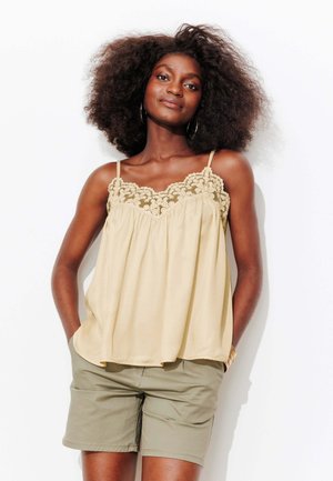 Camisole beige clair avec des détails en dentelle le long du décolleté, associée à un short vert olive. Le tissu semble doux et tombe de manière fluide.