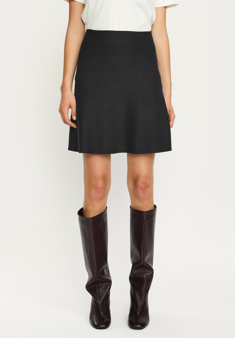 Kobieta ubrana w czarną spódnicę do kolan, biały top z krótkim rękawem i ciemnobrązowe wysokie, skórzane kozaki, stojąca na białym tle.