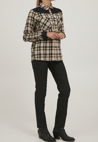 Svart och beige rutigt flanellskjorta med svart krage och manschetter, med en knäppning framtill och två bröstfickor. Kombinerad med svarta byxor.