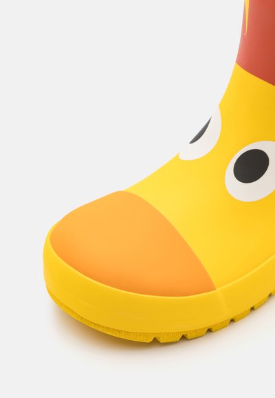 Keltainen kumisaapas, jossa on pyöristetty varvas, suurilla silmäsuunnittelulla ja oranssilla yksityiskohdalla varpaassa. Kuviollinen pohja pidon parantamiseksi.