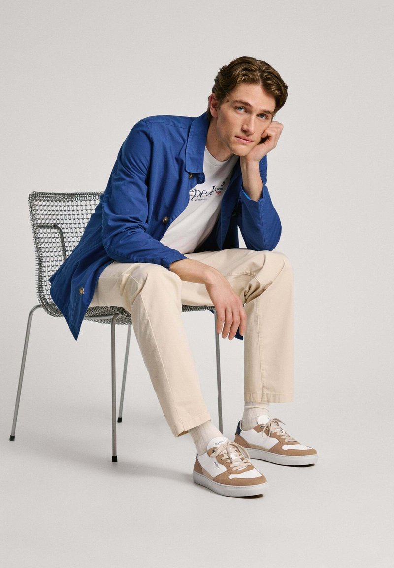 Jachetă albastră, tricou alb cu logo, pantaloni bej și adidași cu nuanțe de maro. Model așezat pe un scaun din sârmă. Fundal minimalist.