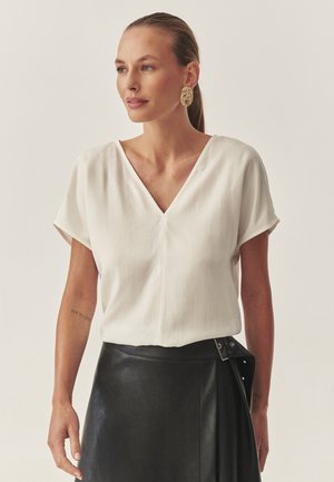 Weißes V-Ausschnitt-Bluse mit kurzen Ärmeln, aus weichem Stoff gefertigt. Kombiniert mit einem schwarzen Lederrock mit Knopf-Akzenten und einer glatten Textur.
