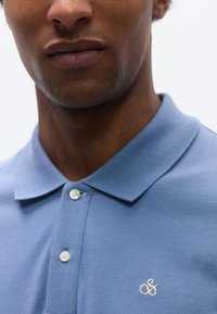 Ανοιχτό μπλε πόλο μπλούζα από υφασμάτινη υφή, με τρία κουμπιά και λευκό κεντημένο λογότυπο στο αριστερό στήθος.