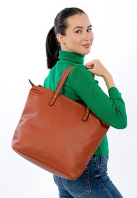 Brown leather tote bag with textured surface, dual straps, and a zippered closure. Model poses with bag on shoulder against white background.